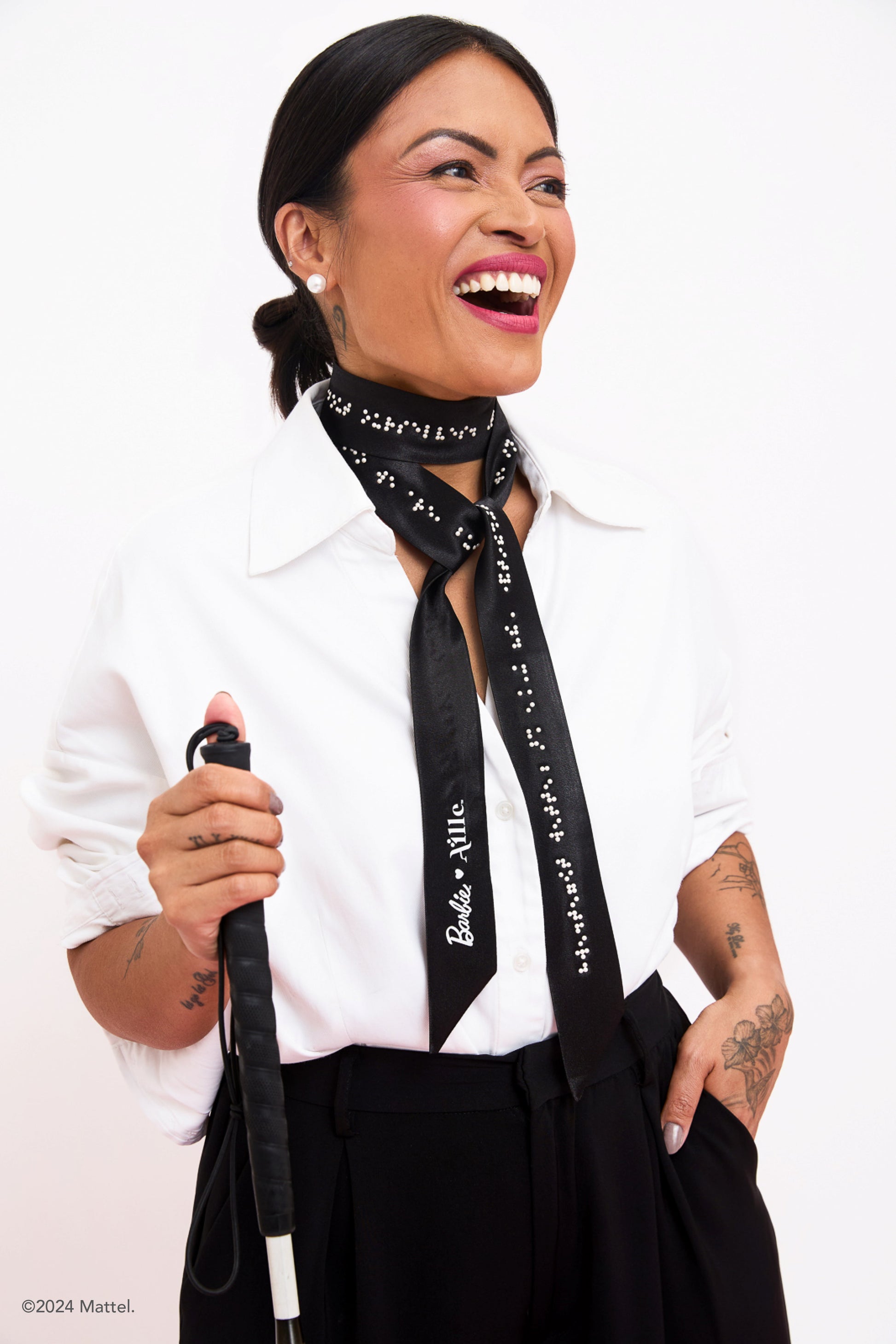 Blind woman with white cane smiles while wearing thin black scarf accessory with white braille beadwork and the Barbie x Aille logo.
