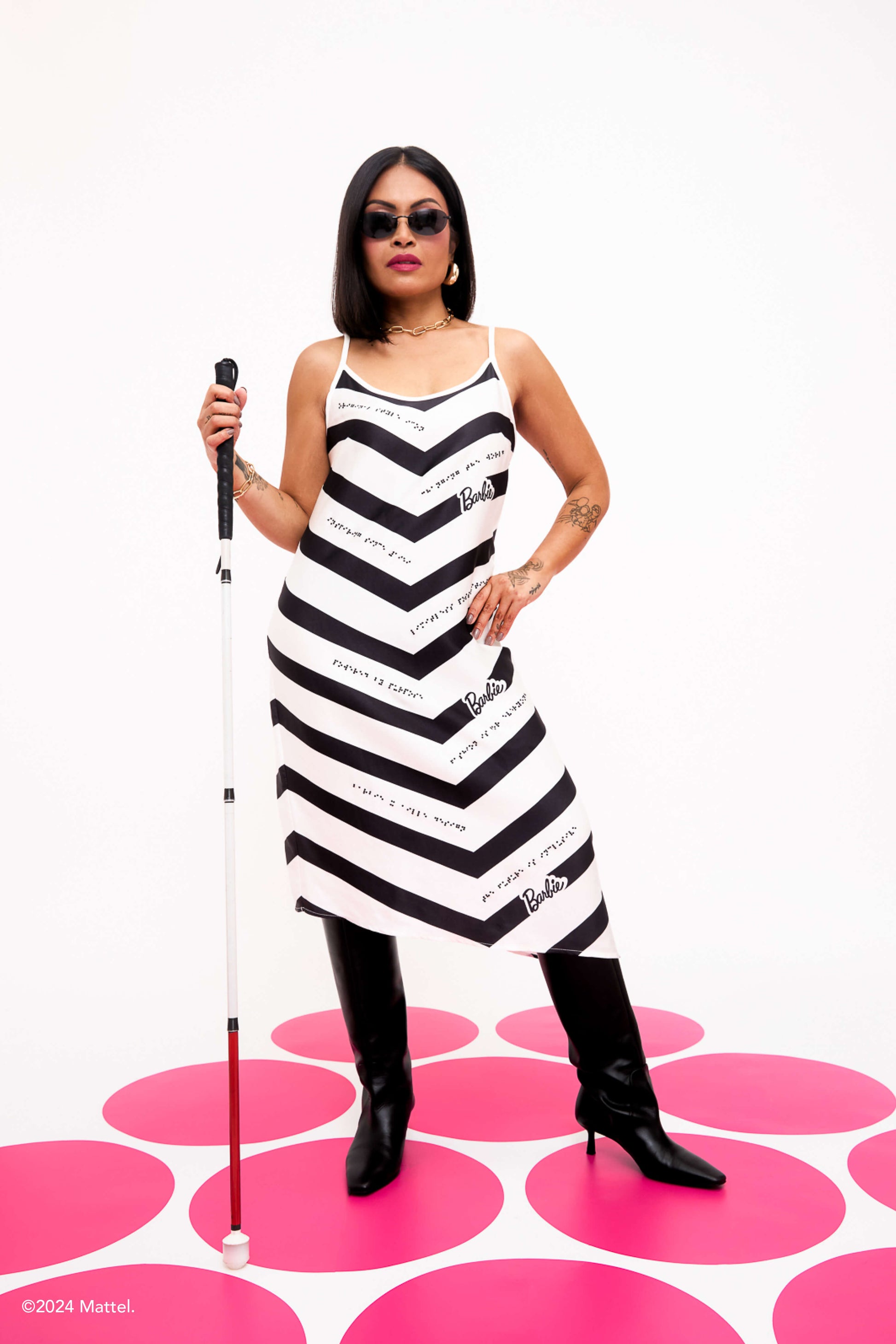 Blind woman with dark hair and sunglasses stands confidently, holding a white cane, wearing a black and white chevron braille dress.