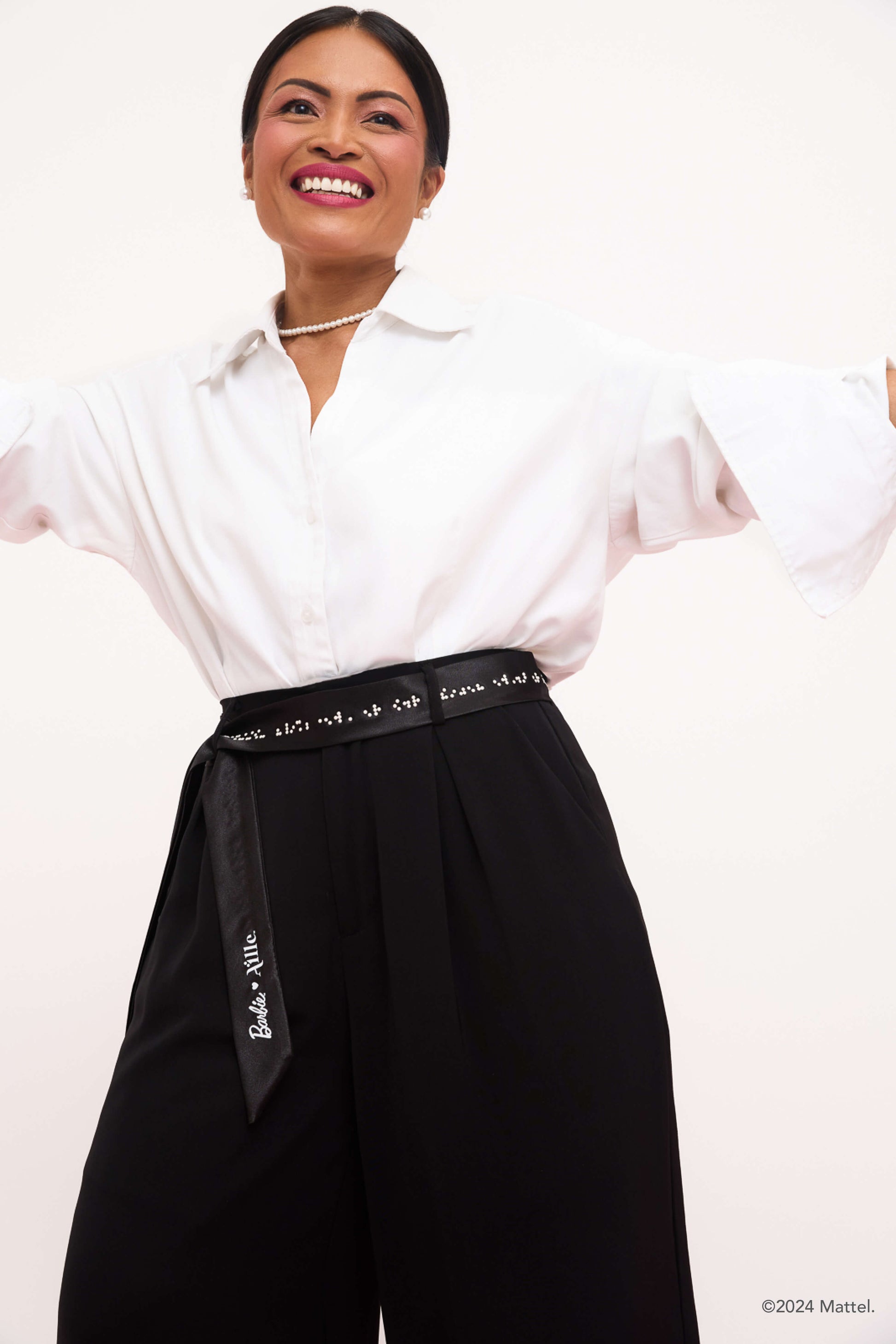 Woman smiling wearing white collared shirt and black trousers. A black belt with white braille is tied around her waist as a belt.