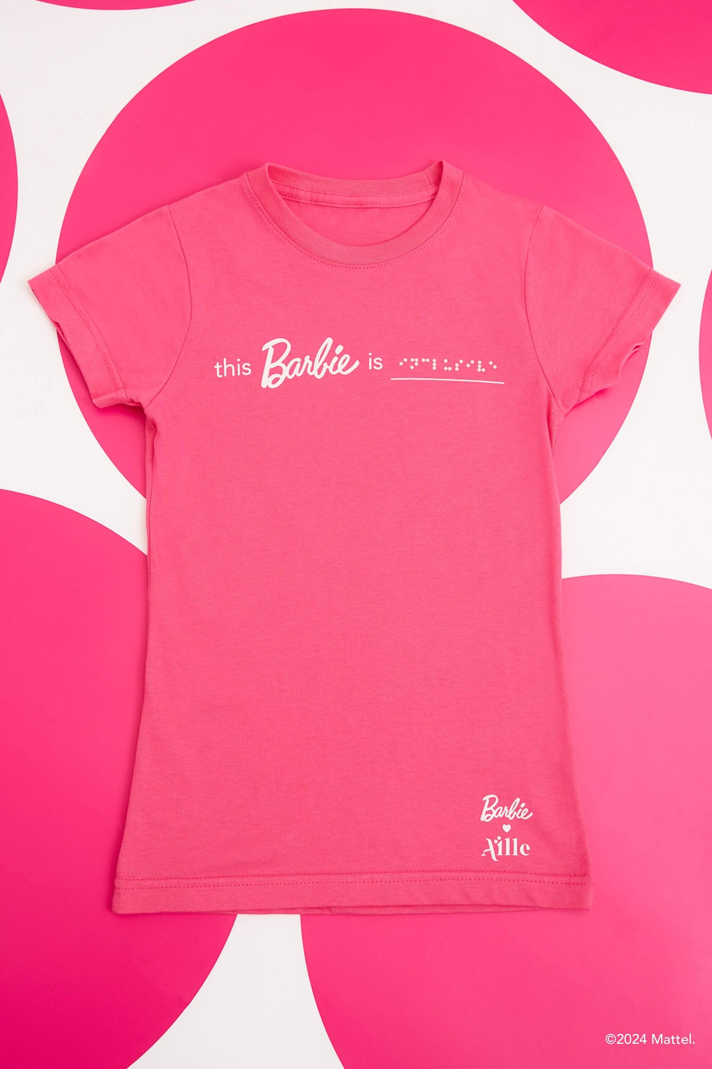 Flat lay photo of a bright pink T-shirt with white braille text and braille on a pink polka dot background,