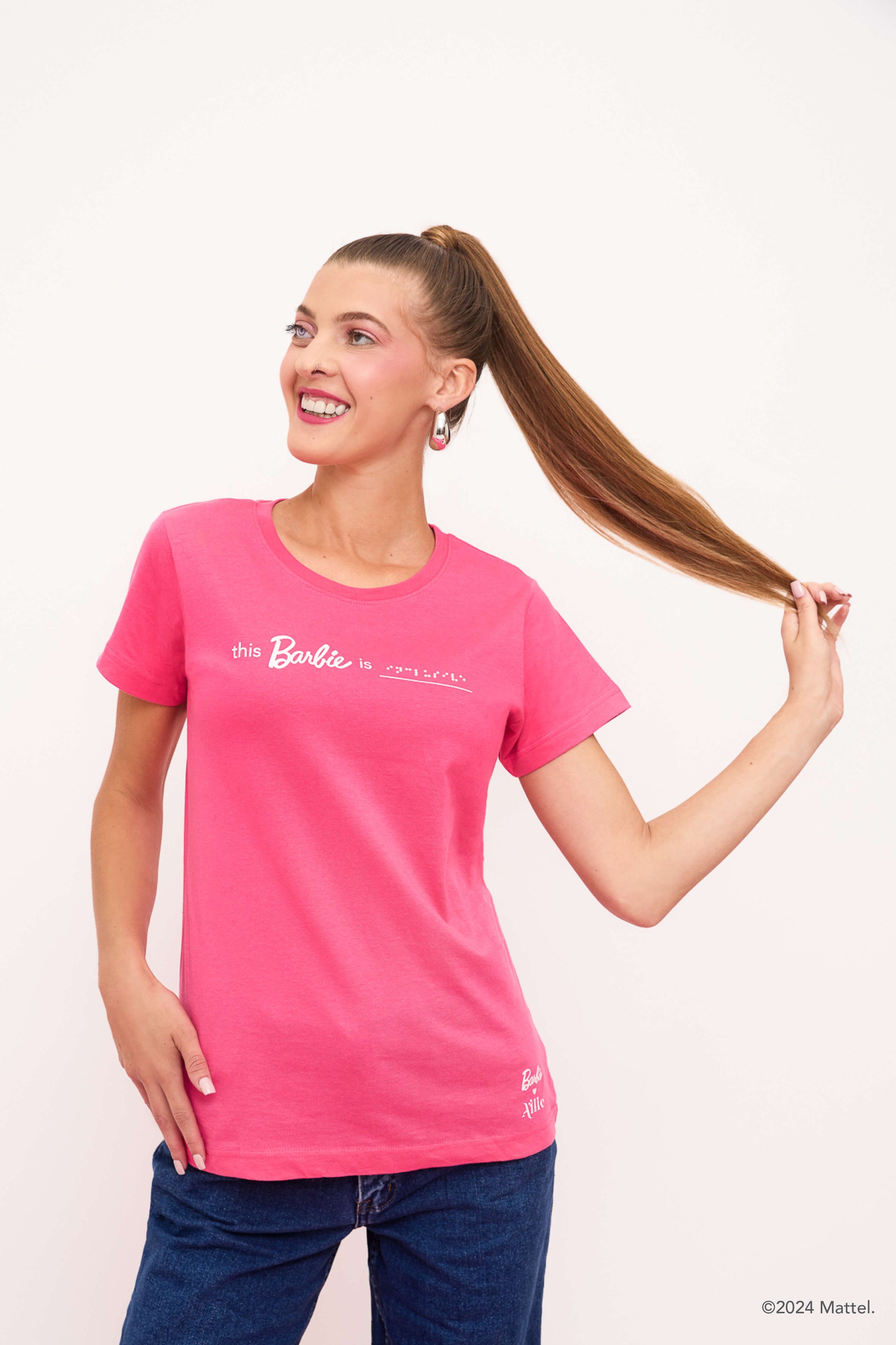 Blind woman playfully holding her ponytail, wearing a bright pink T-shirt and jeans, smiling brightly.
