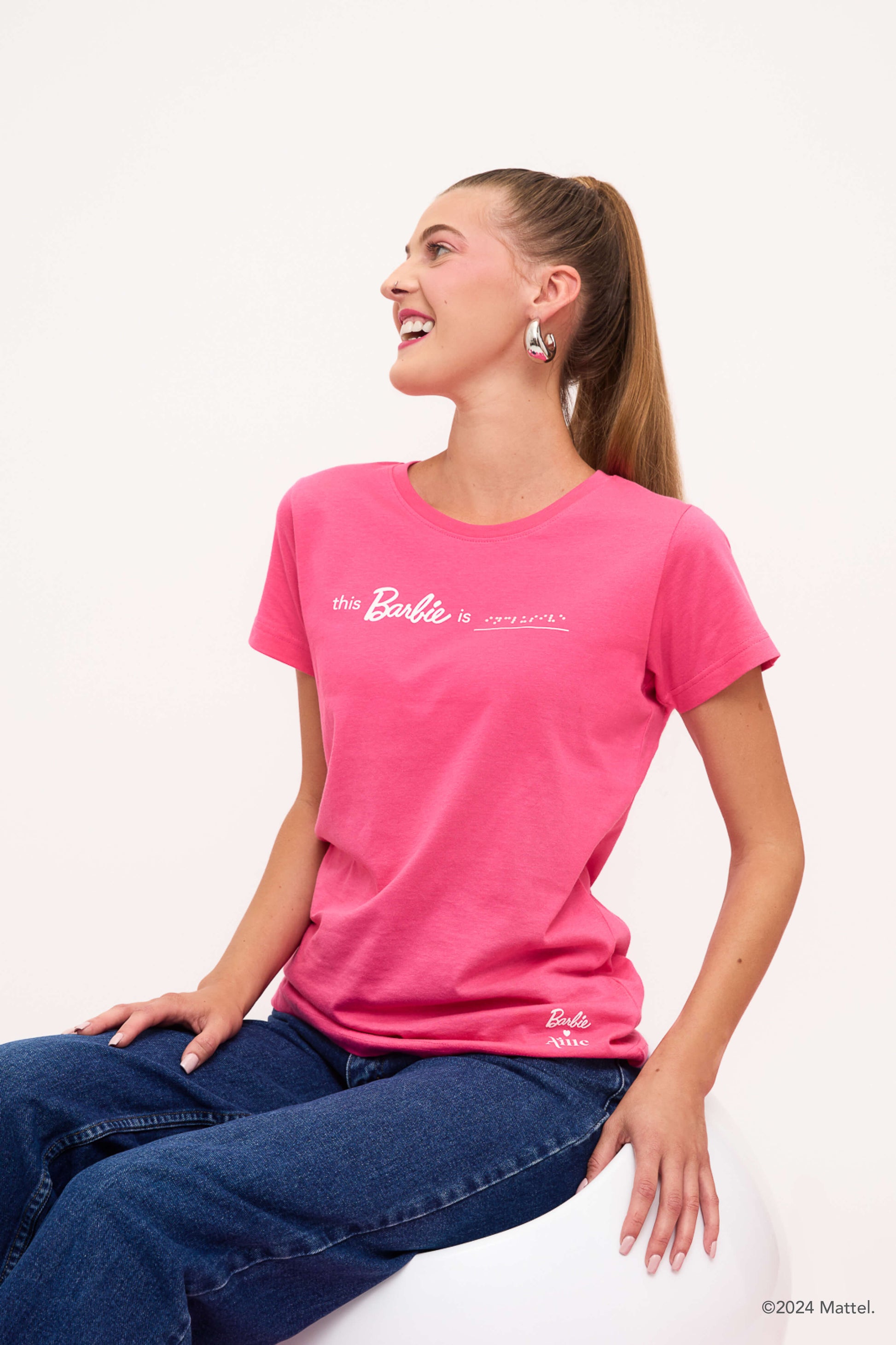 A blind woman sitting and smiling wearing a bright pink T-shirt and jeans.