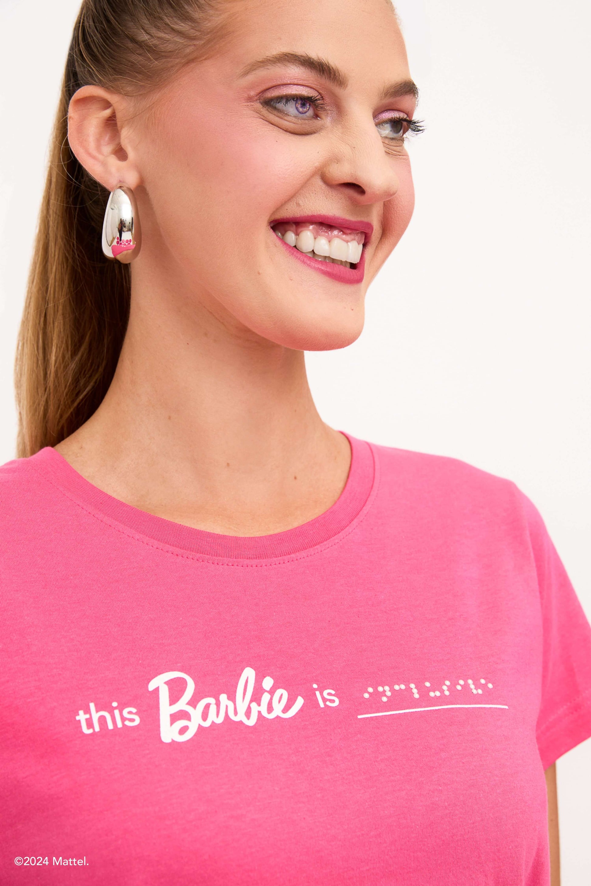 Close up photo of blind model showcasing a beautiful prosthetic eye. Her bright pink T-shirt features white text and braille that reads "This Barbie is Inclusive".