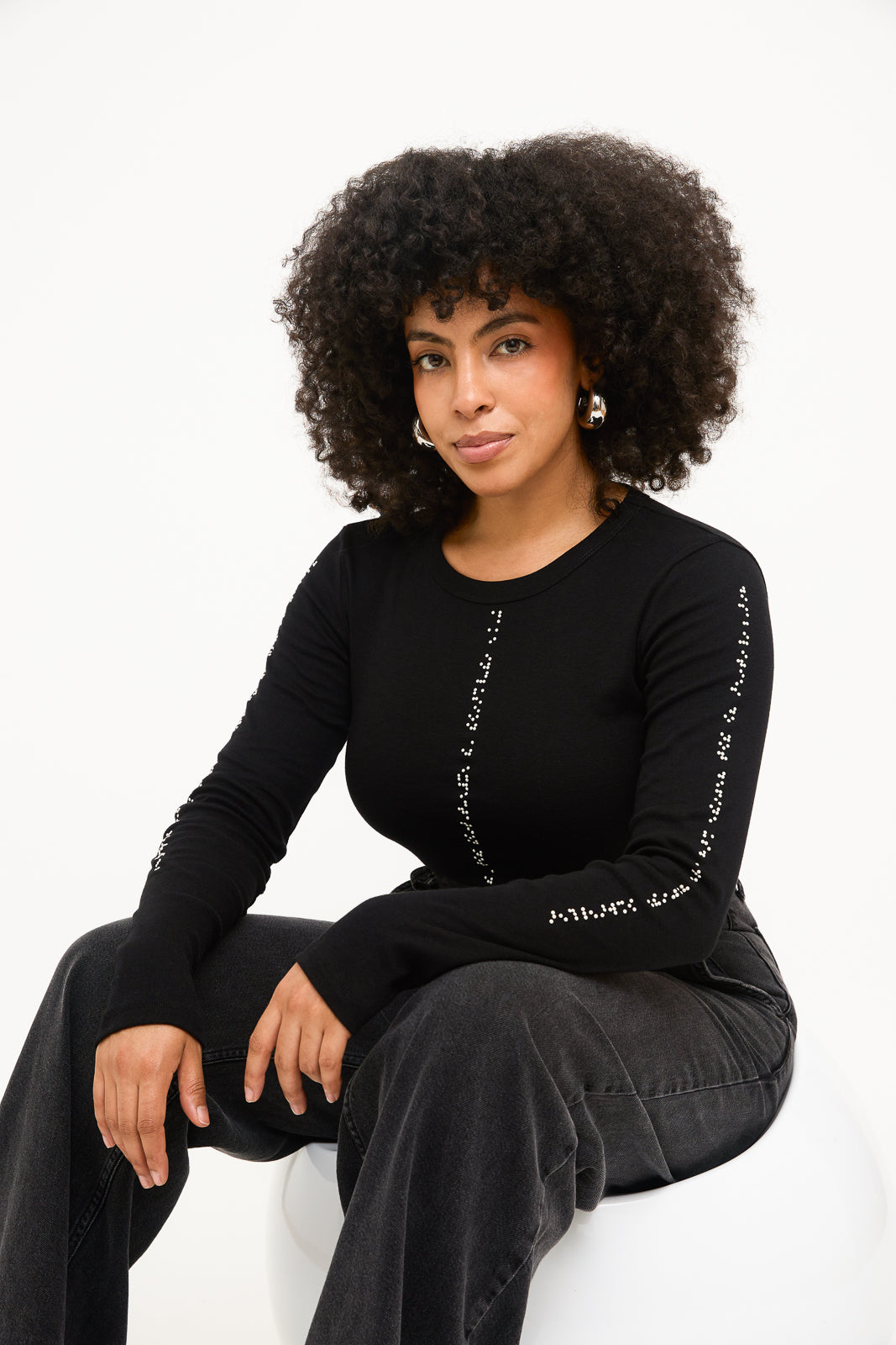 Model is seated on a white chair, her arms resting on her knee to showcase the long lines of white braille on her black long sleeve shirt.