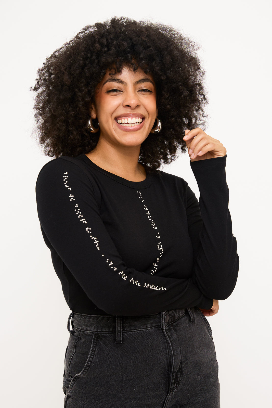 Model's hand is resting on her hip, and she is smiling with her curly hair framing her face. This view shows the white braille on the opposite sleeve of her black shirt.