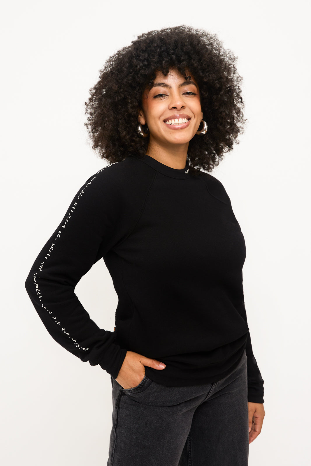 Model standing with a hand in her pocket, wearing a black crewneck sweater with white braille beadwork running down the right sleeve. The look is paired with high-waisted black jeans.