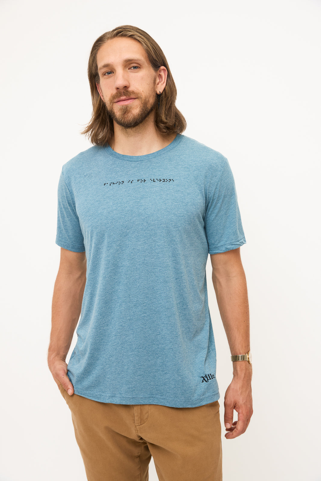 Front View of Blue GreyT-Shirt: Young man standing, hand in pocket, smiling and looking at camera. He's wearing a blue grey t-shirt with black braille beadwork on the chest.