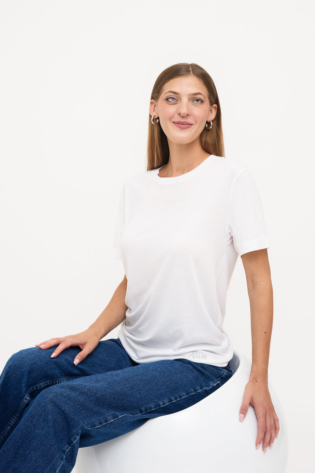 Seated in Woman's White T-Shirt: Young blind woman seated on a white chair, looking at the camera, wearing a white t-shirt with white braille beadwork below the neckline.