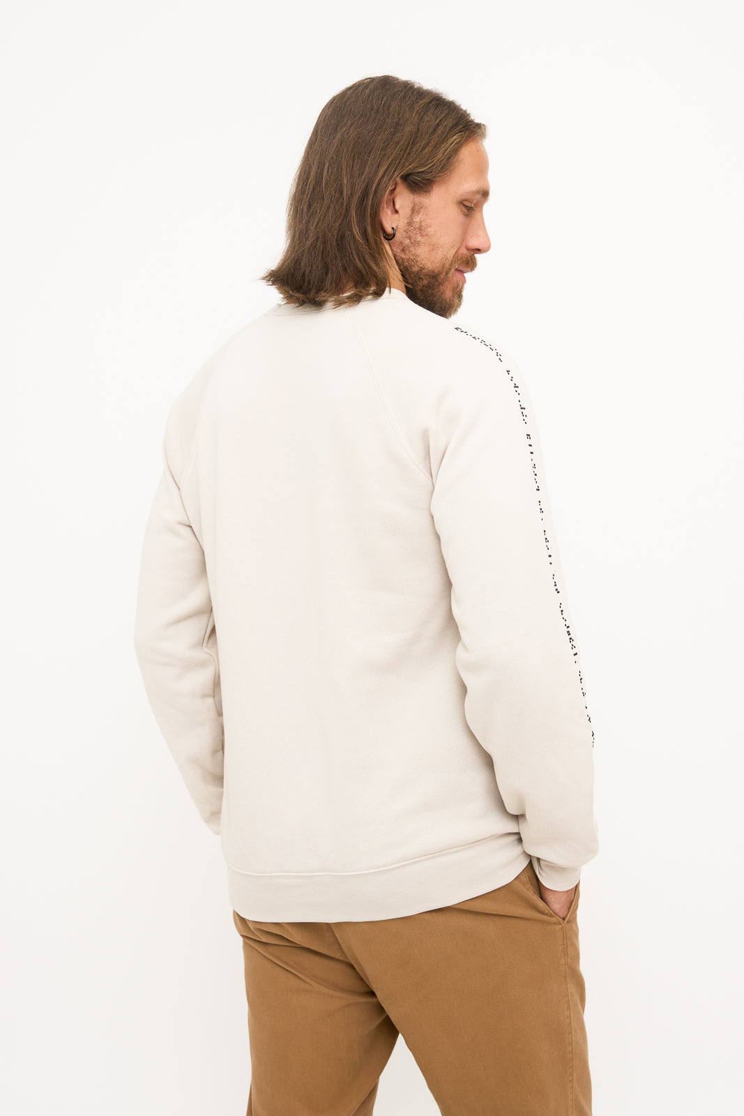 Model facing away from the camera at a slight angle. This view shows the plain back of the beige sweater and emphasizes the black braille running down the right sleeve.