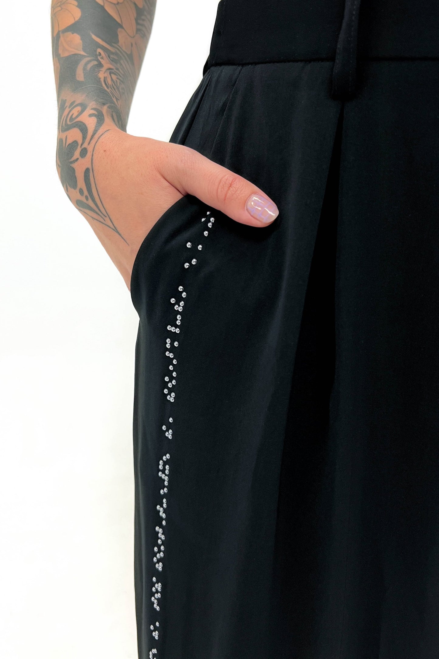 A close-up of the side seam pocket area of the black wide-leg pants, showing the detailed chrome braille beadwork. The model's hand is casually placed in the pocket.