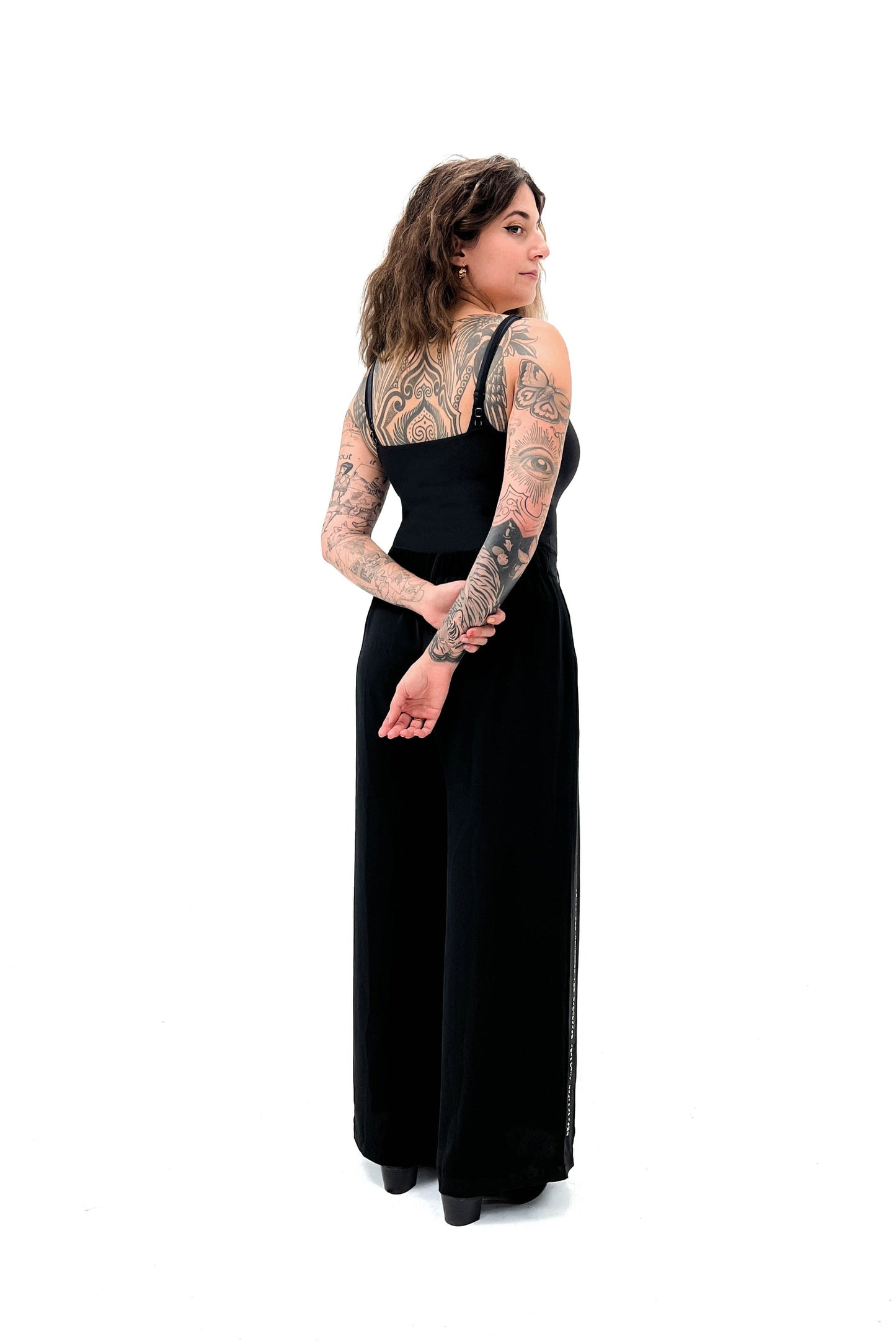 Full body back view of black wide-leg pants. Model's hands are behind her back. She has beautiful tattoos along her back and arms.