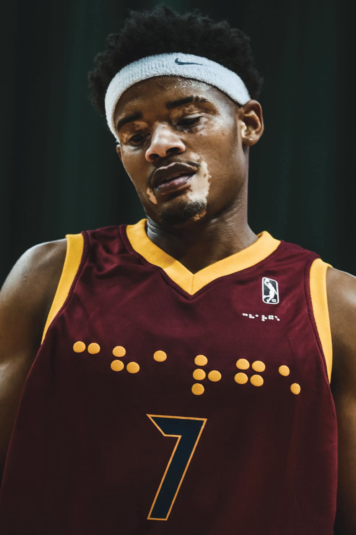 Close up photo of an NBA player with vitiligo. He is wearing a custom braille beaded jersey and a white Nike sweatband. The jersey is maroon with gold accents and the braille beadwork is white.