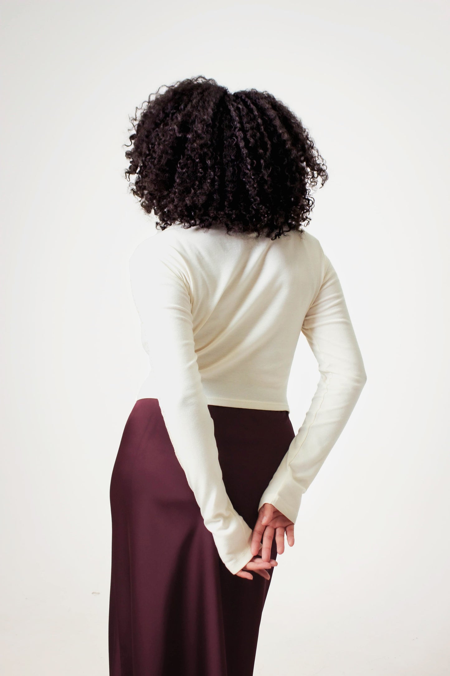 Back view of cropped cream long sleeve shirt. Model's hands are behind her back to showcase length of sleeves. Shirt is paired with maroon satin slip skirt.