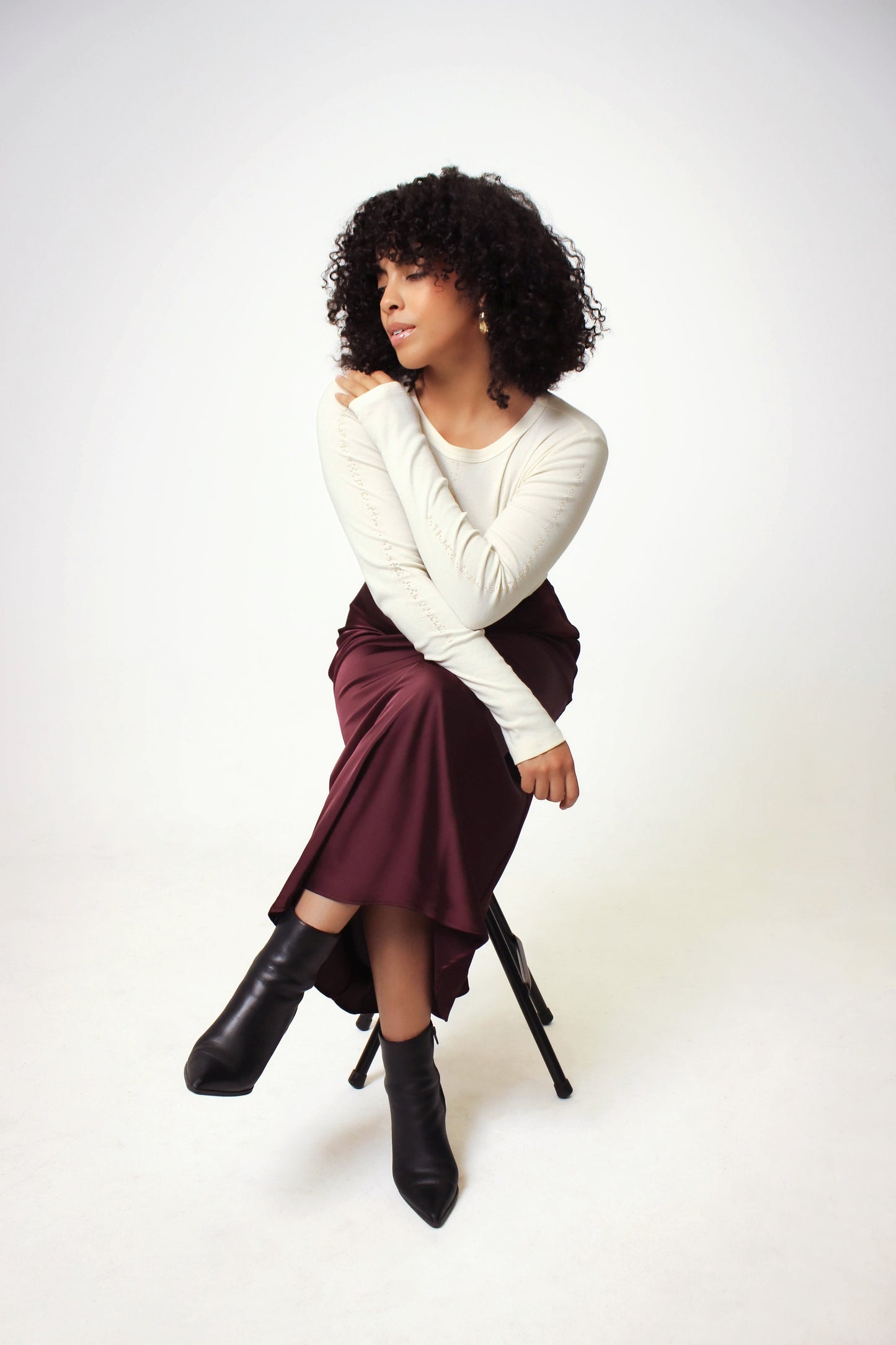 Model is seated wearing the cropped cream long sleeve shirt paired with a maroon slip skirt and black boots. The model is posing with her arms to showcase the gold braille beadwork on her sleeves.