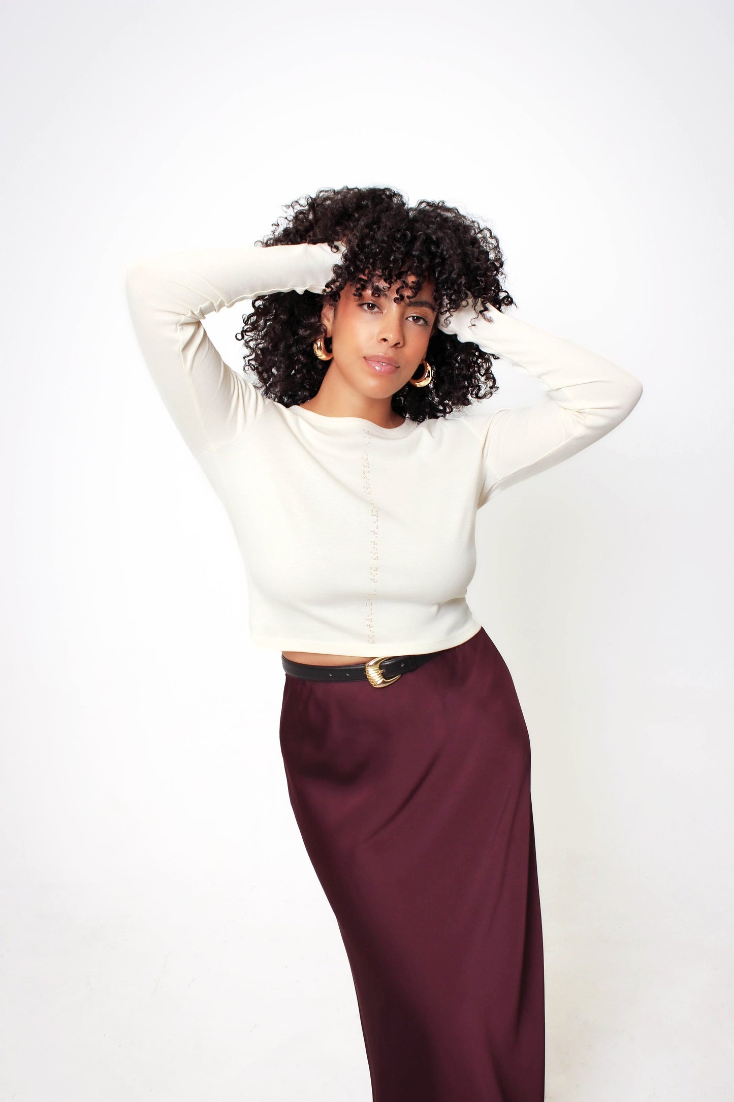 Front view of cropped cream long sleeve shirt. Model's hands are on her head to showcase cropped length of shirt. Shirt is paired with maroon satin slip skirt and black and gold belt.