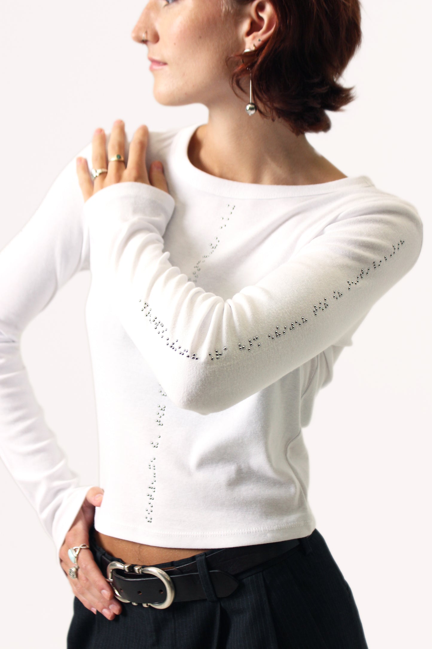 Model's left hand is on her shoulder, showcasing the beautiful chrome beadwork on her white long sleeve shirt. The model accessorized the look with black pants, a belt, and silver jewelery.