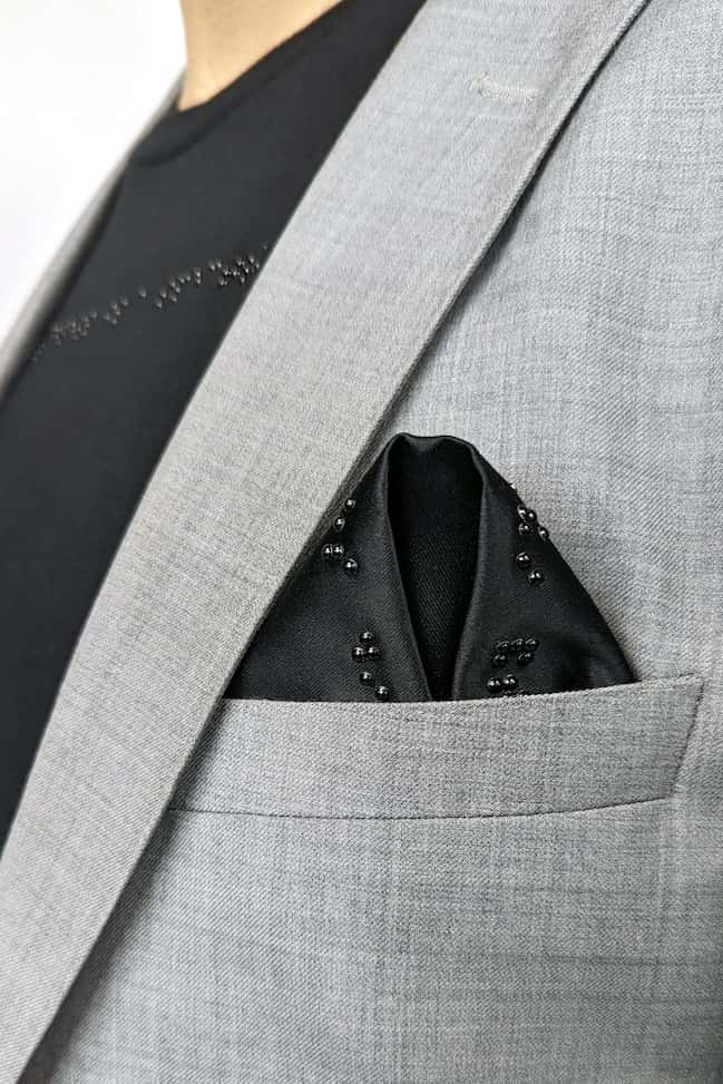 Close up of black folded pocket square in light grey suit. Man is also wearing black t-shirt with black braille beadwork.