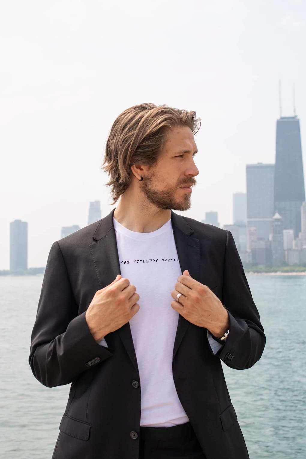 White man with long hair and beard wearing white braille shirt and black blazer