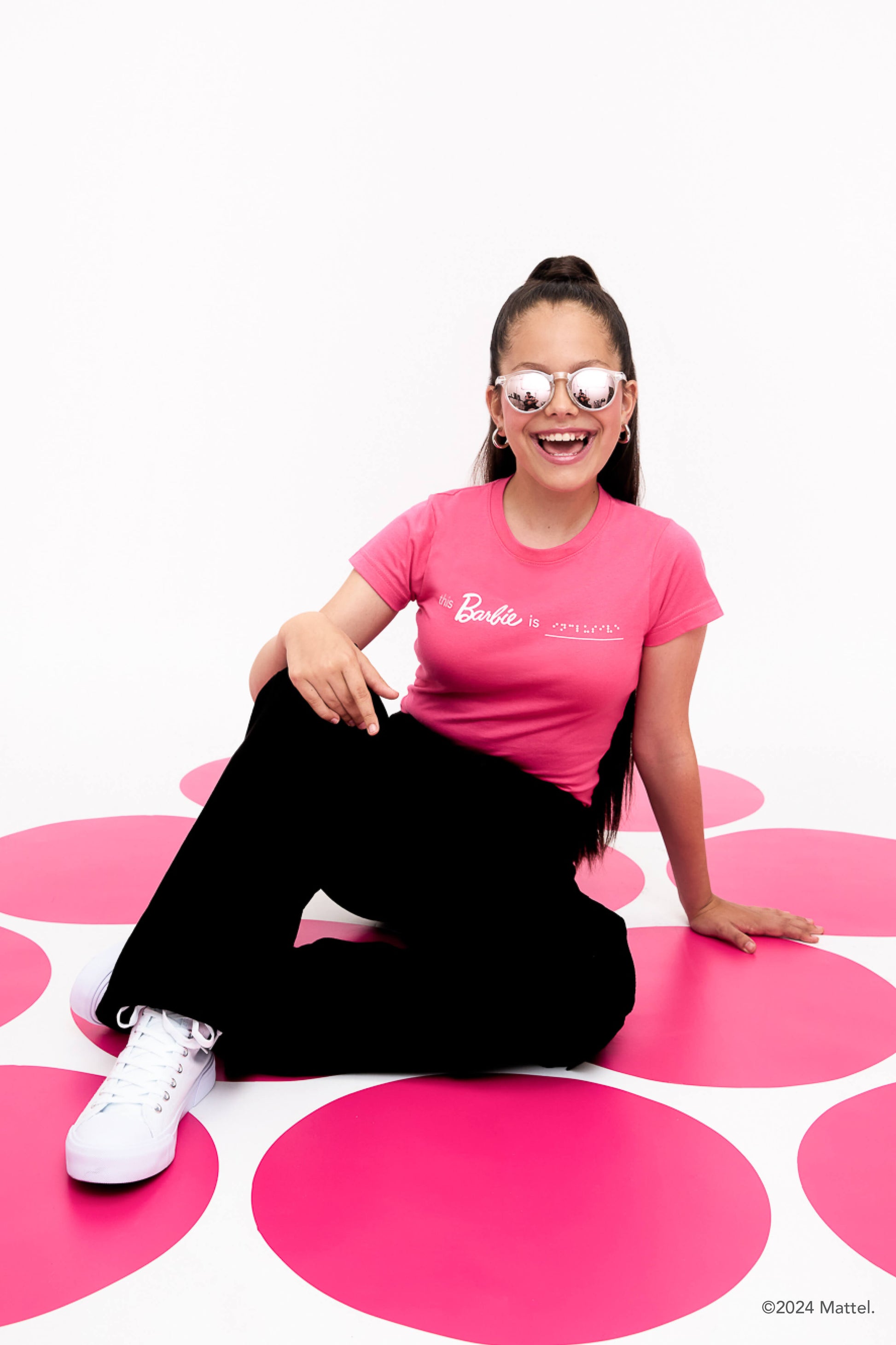 Young girl sits on a pink polka dot floor, laughing and. She is wearing a bright pink T-shirt with white braille, black cargo pants, and white sneakers. 