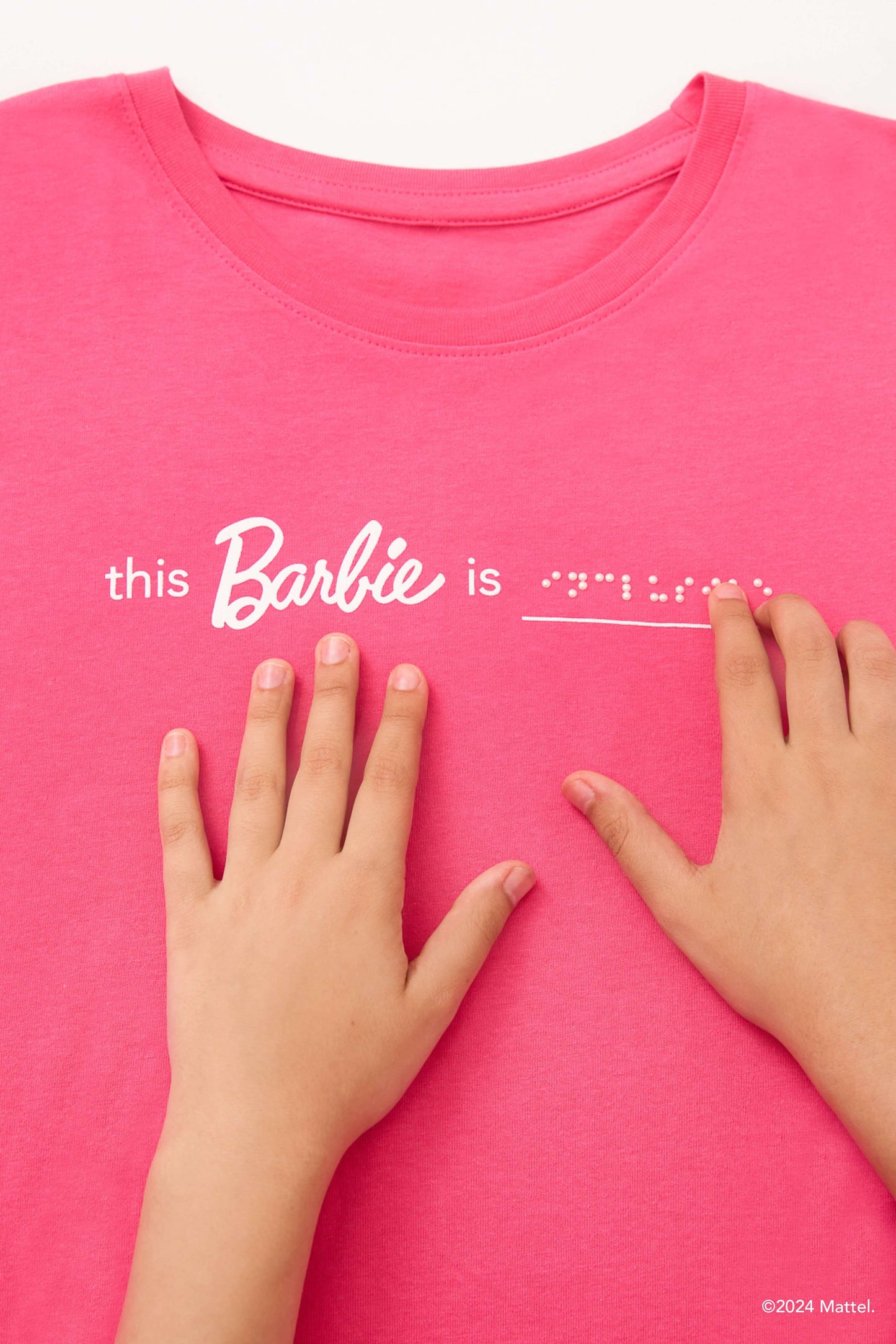 Close-up of a pair of young girl's hands reading braille on a bright pink T-shirt that says "this Barbie is" in print and "inclusive" in white braille beadwork.