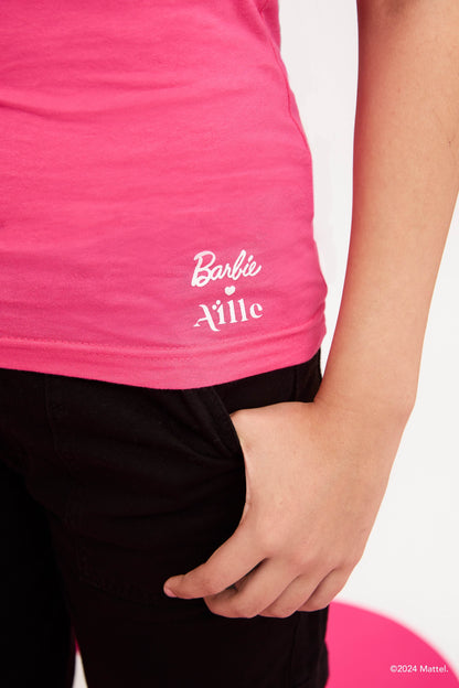 Close-up of young girl wearing a bright pink T-shirt with the "Barbie x Aille" logo in white near the hem. Her hand is resting in her pant pocket.