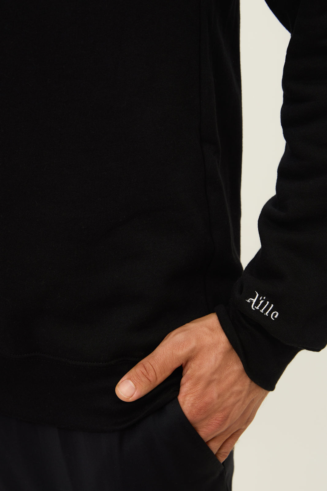 Close up of Aille Design logo embroidered in white on the cuff of the left sweater sleeve.