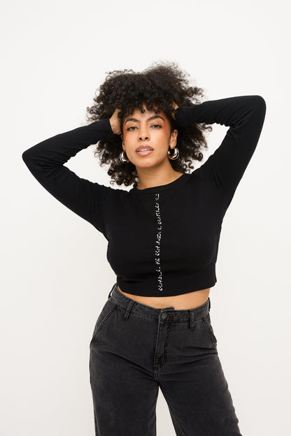 Model standing with her hands in her curly hair, wearing a black cropped long-sleeve shirt with white braille beadwork running vertically down the center front and both arm sleeves. The look is paired with high-waisted black jeans.