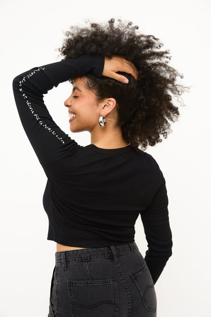 Strong pose of model standing in a side profile, smiling with one hand in her curly hair. This view showcases plain black back of shirt, and white braille on raised sleeve.