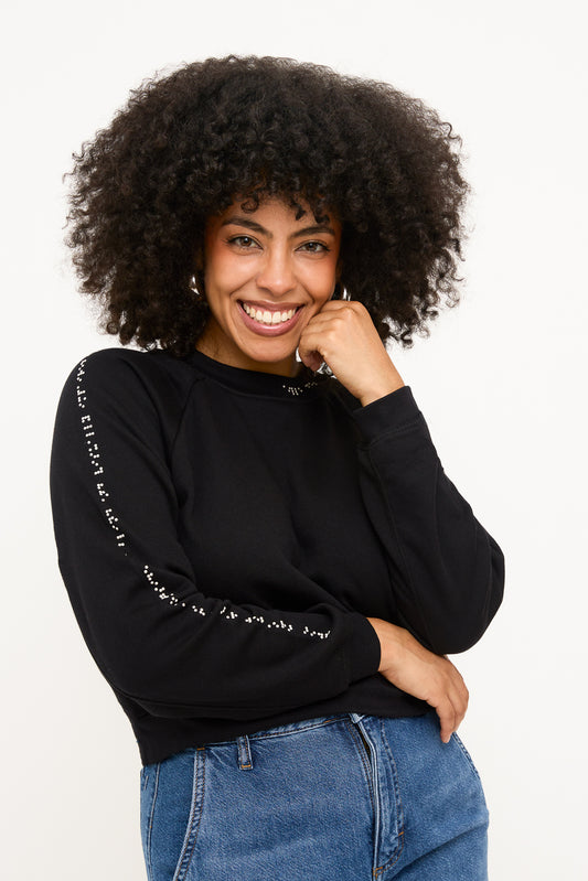 Model standing with a hand on her hip and a hand on her chin, wearing a black cropped sweater with white braille beadwork running down the right sleeve. The look is paired with high-waisted blue jeans.