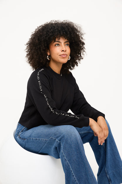 Model is seated on a white chair, her arms resting on her knee to showcase the long line of white braille on her right sweater sleeve.