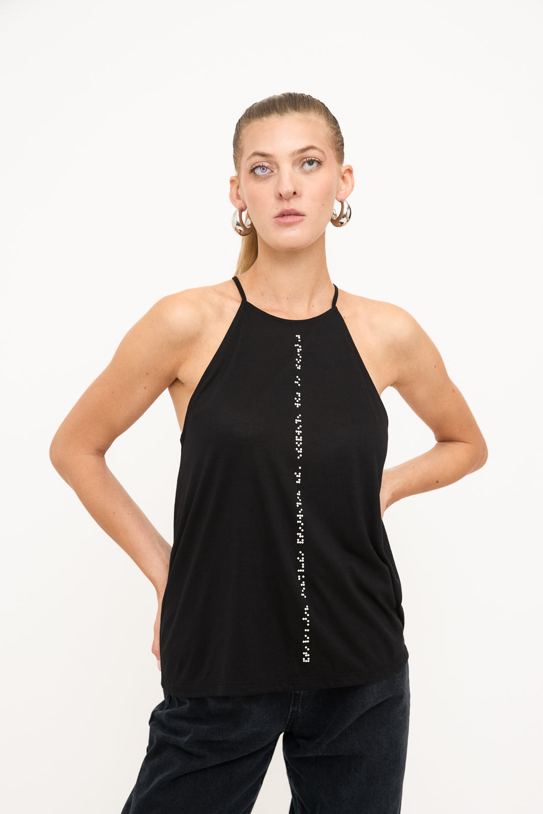 Blind model poses confidently with hands on hips, wearing a black high neck tank top with white braille beadwork down the front. The look is paired with dark black jeans.