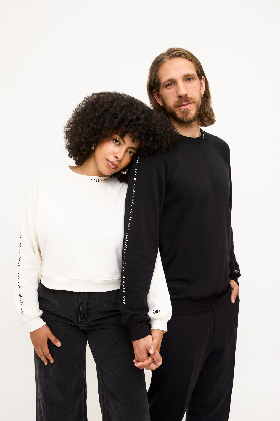 Holding hands, a women wearing a white cropped sweater with black braille beads rests her head on a man wearing a black crewneck sweater with white braille beads.