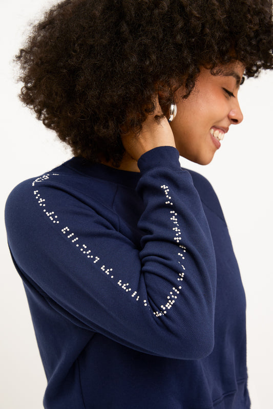 A women wearing a navy blue cropped sweater with white braille beads has her hand in her hair, emphasizing the braille along her right sleeve.