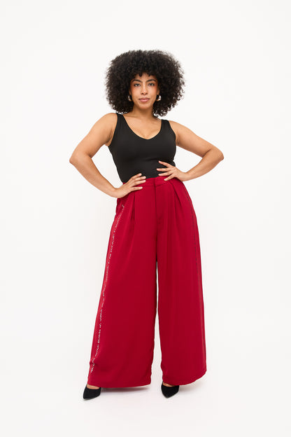 Model poses confidently with hands on hip. She is wearing red wide-leg pants with black and white braille beadwork along the pant leg side seam. The pants are styled with a black sleeveless top.