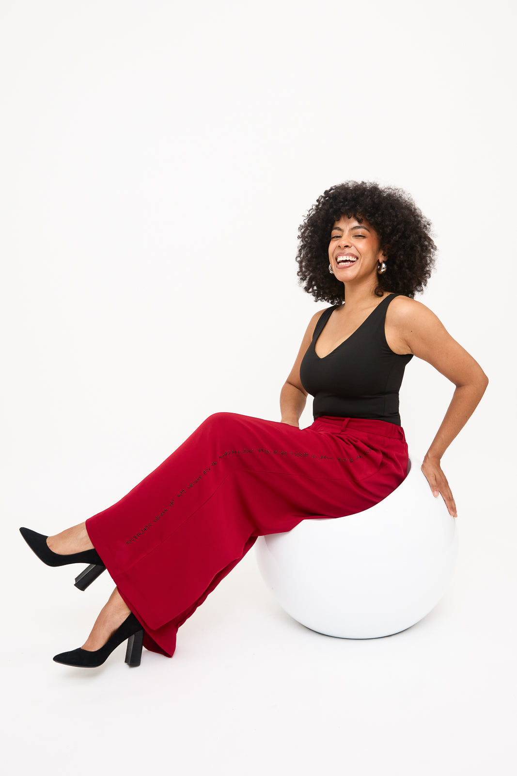 Fully body photo of seated model wearing red wide-leg pants showcasing the black braille along one pant leg.