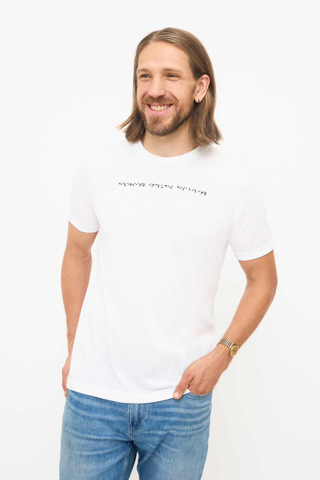 Front View of White T-Shirt: Young man standing, hand in pocket, smiling and looking away. He's wearing a white t-shirt with black braille beadwork on the chest.