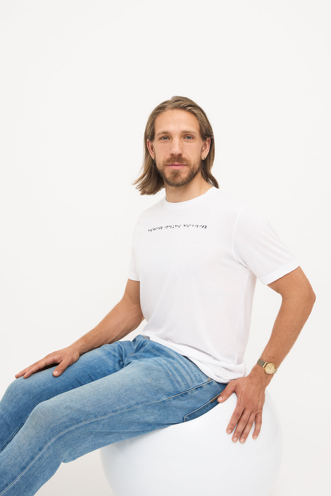 Seated in White T-Shirt: Young man seated in a white chair, wearing a white t-shirt with black braille beadwork on the chest.