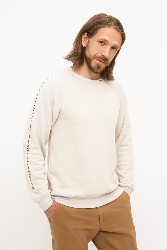 Young man standing, hands in pockets, smiling and looking at camera. He's wearing a beige crewneck sweater with black braille beadwork on the right sleeve and collar.