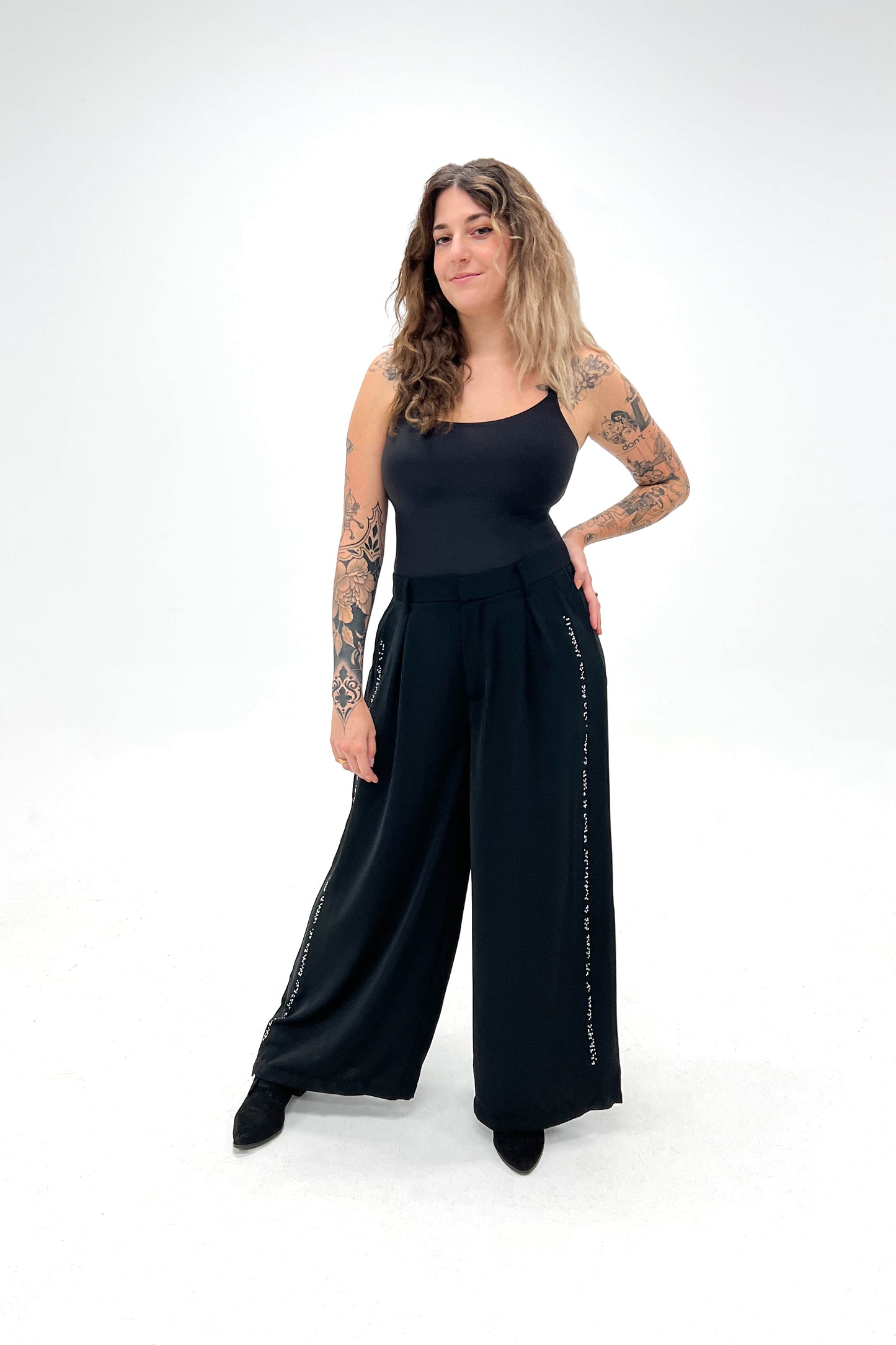 Model poses with one hand on her hip. She is wearing black wide-leg pants with chrome braille beadwork along the pant leg side seam. The pants are styled with a black sleeveless top.