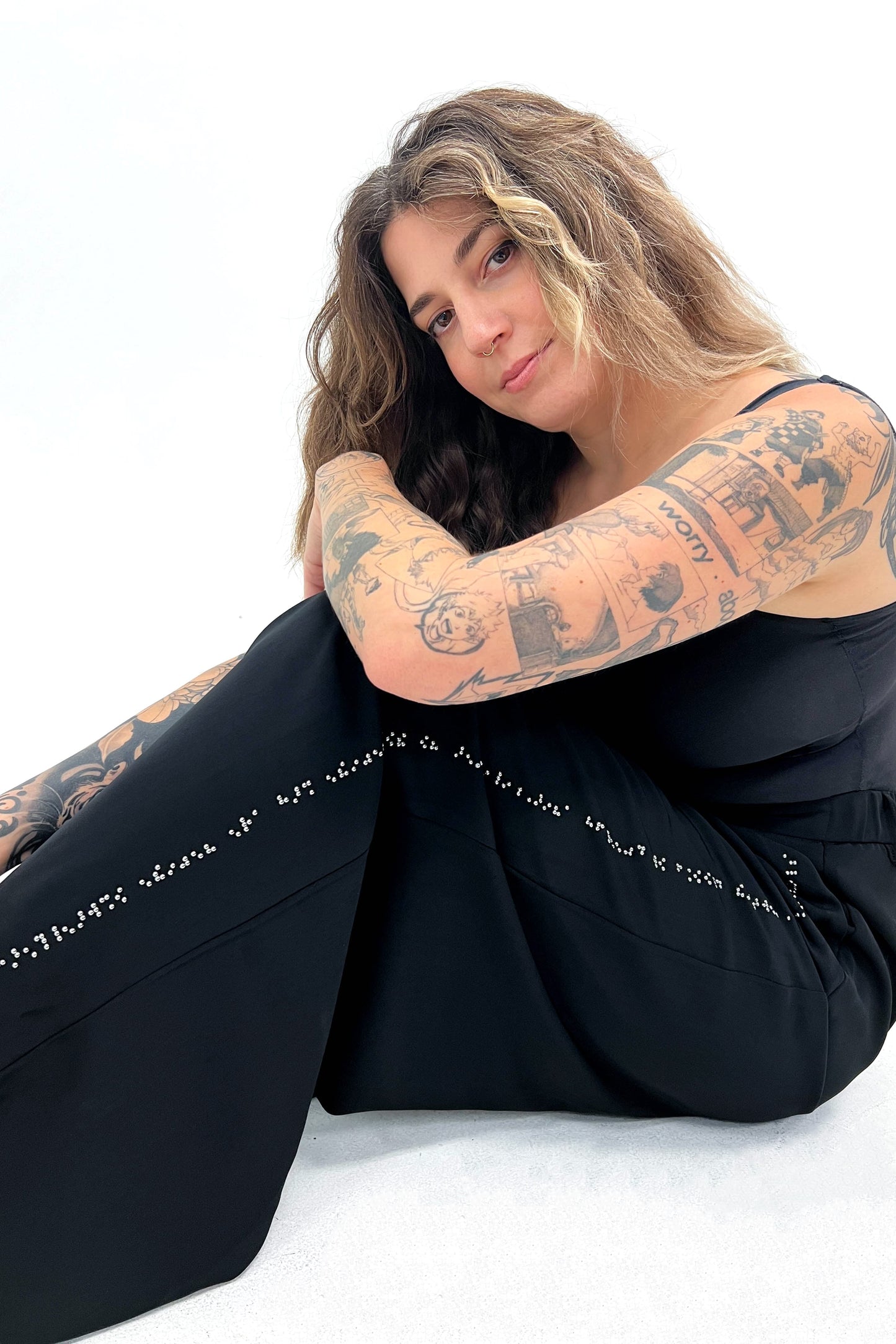 Close up photo of a woman with tattoos on her arms. She's seated with her arm resting on her knee. She's wearing black pants adorned with chrome tactile braille beadwork, which runs alongside the side seam of the pant which is featured prominently in this pose. Her detailed tattoo art complements the intricate braille beadwork on the pants, creating a visually striking contrast.