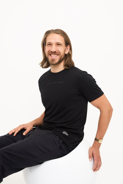 Seated in Black T-Shirt: Young man seated in a white chair, smiling, wearing a black t-shirt with black braille beadwork on the chest.