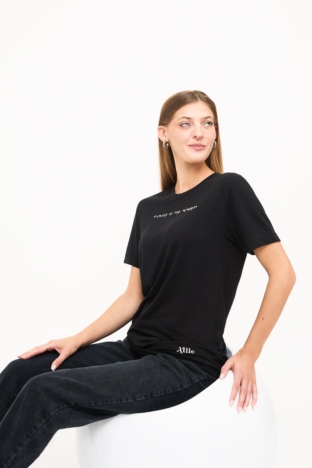 Seated in Woman's Black T-Shirt: Young blind woman seated, looking to the side, wearing a black t-shirt with white braille beadwork below the neckline.