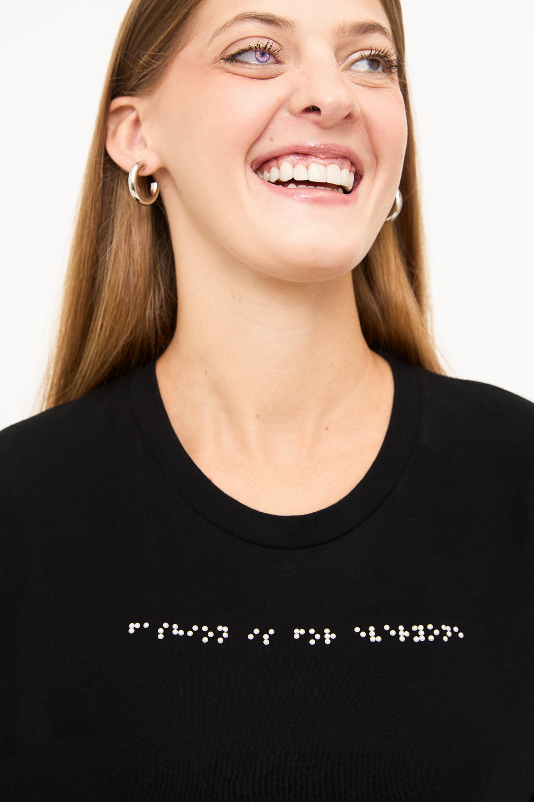 Blind model with beautiful prosthetic eye and a big smile and wears black T-shirt with white braille beadwork that reads "fashion is for everyone".