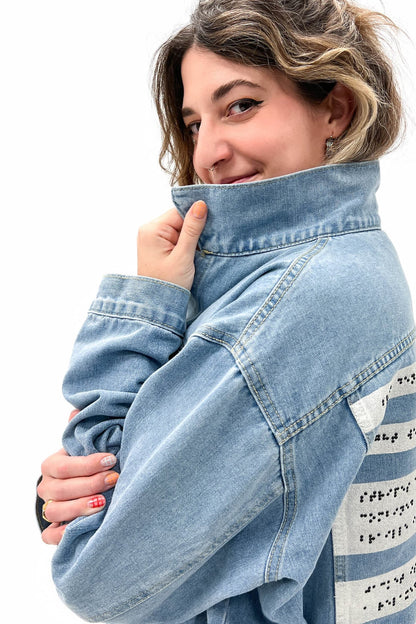A close-up shot of a smiling woman pulling the collar of their light denim jacket toward their face, exuding a playful and confident vibe. The braille panel on the back is partially visible.