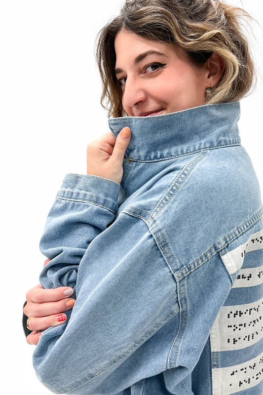 A close-up shot of a smiling woman pulling the collar of their light denim jacket toward their face, exuding a playful and confident vibe. The braille panel on the back is partially visible.