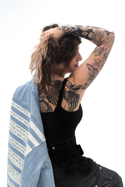 A woman with intricate tattoos on their arms and shoulder poses in profile, with one arm raised and hand resting on the back of their head. They are wearing a black top and holding the light denim jacket over their shoulder, revealing the jacket’s braille panel.