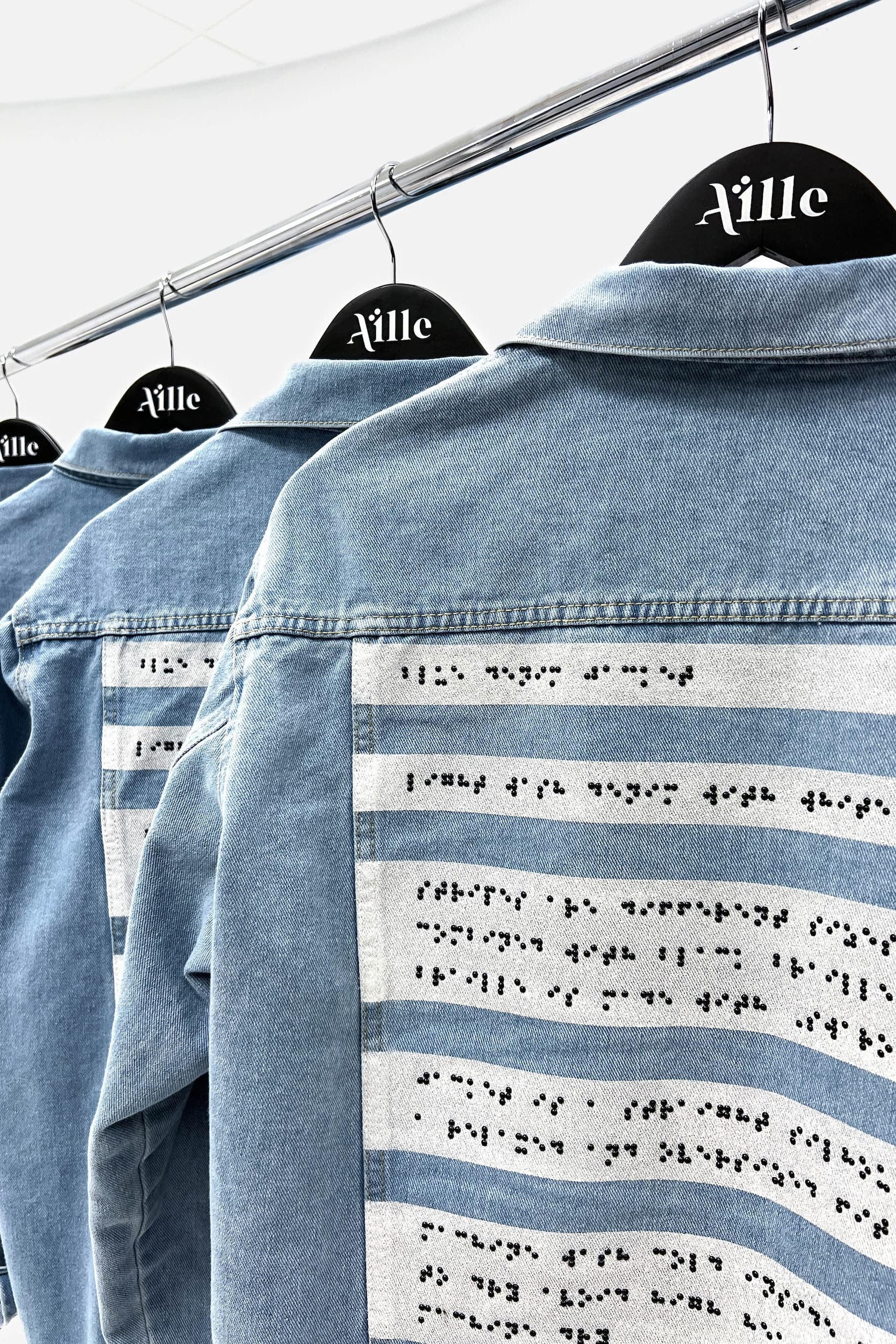  close-up view of multiple light blue denim jackets hanging on a garment rack, each displayed on black hangers with the Aille Design logo in white. The backs of the jackets feature painted white stripes with black braille beading that runs horizontally across each stripe.