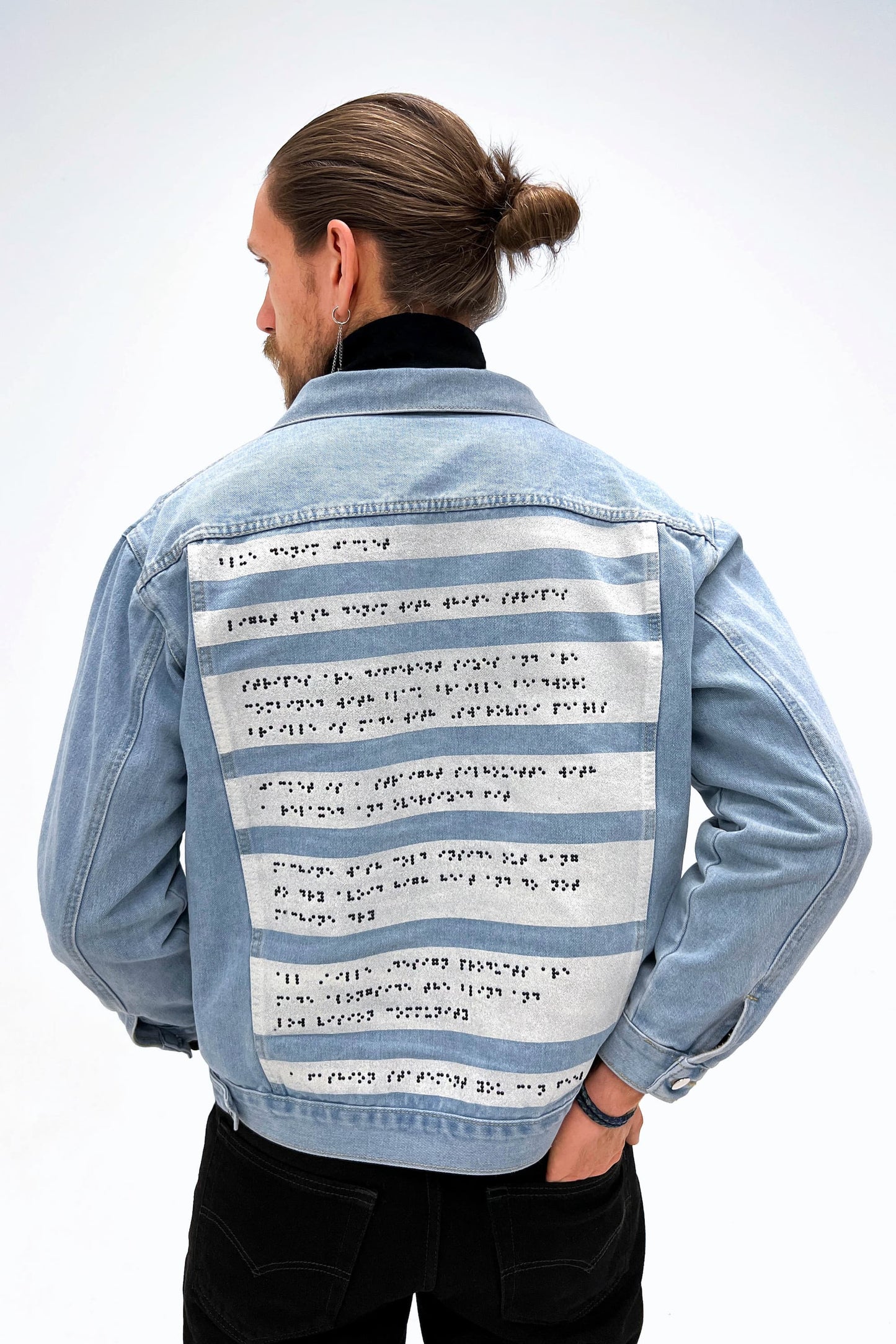 A back view of a man wearing the light wash braille denim jacket. His hair is tied up in a bun, and he is wearing black jeans, a black turtleneck, and a long silver earring. He is standing with one hand in his back pocket.