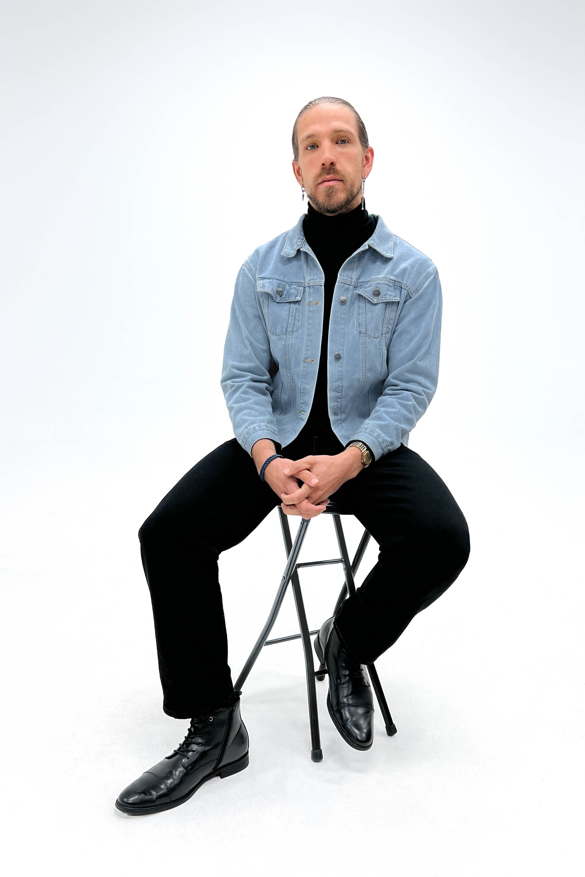 A man sits on a black stool, wearing the light denim jacket with a black turtleneck, black jeans, and black dress boots. Their hands are resting in their lap.