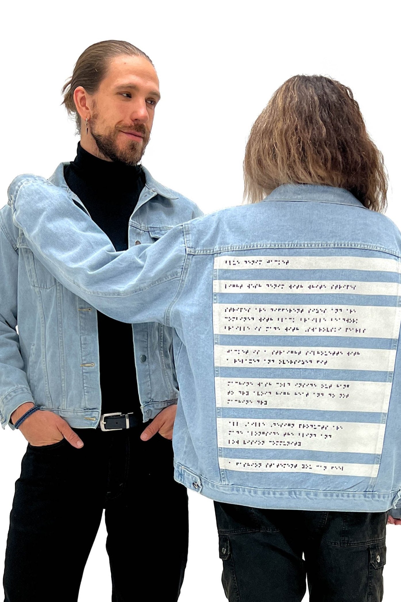 Two people are shown wearing matching light denim jackets, both featuring Aille Design’s signature braille beaded panel on the back. The person on the left, facing forward, wears a black turtleneck underneath their jacket and has a calm expression. The person on the right has their back to the camera, showcasing the detailed braille beadwork. The back of the jackets feature painted white stripes with black braille beading that runs horizontally across each stripe.